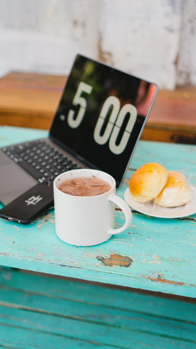 Laptop showing time next to coffee and pastries on table.